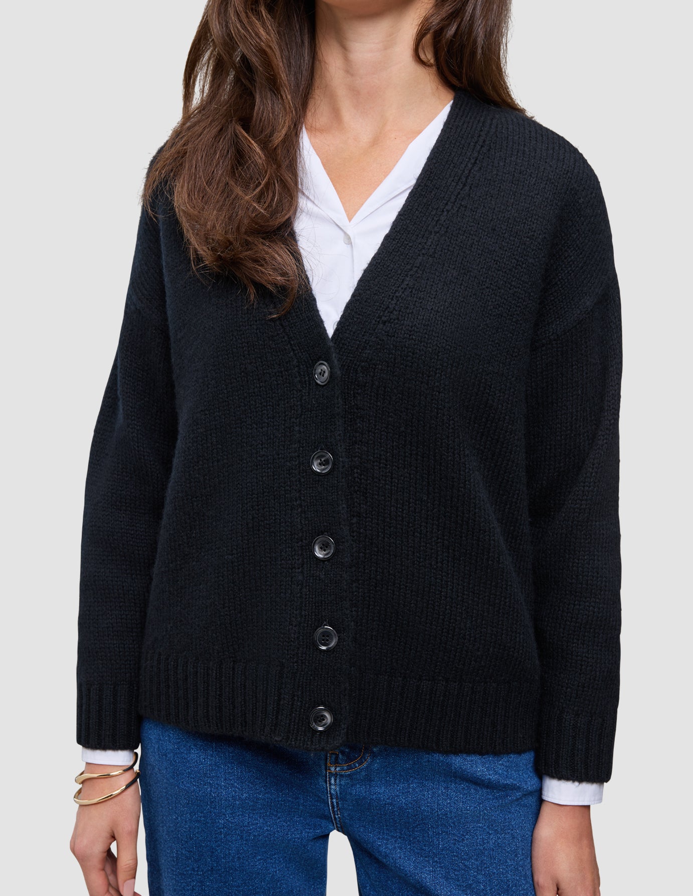 Kvinners tykk, myk ullstrikket cardigan i svart, båret over en hvit skjorte og blå jeans.