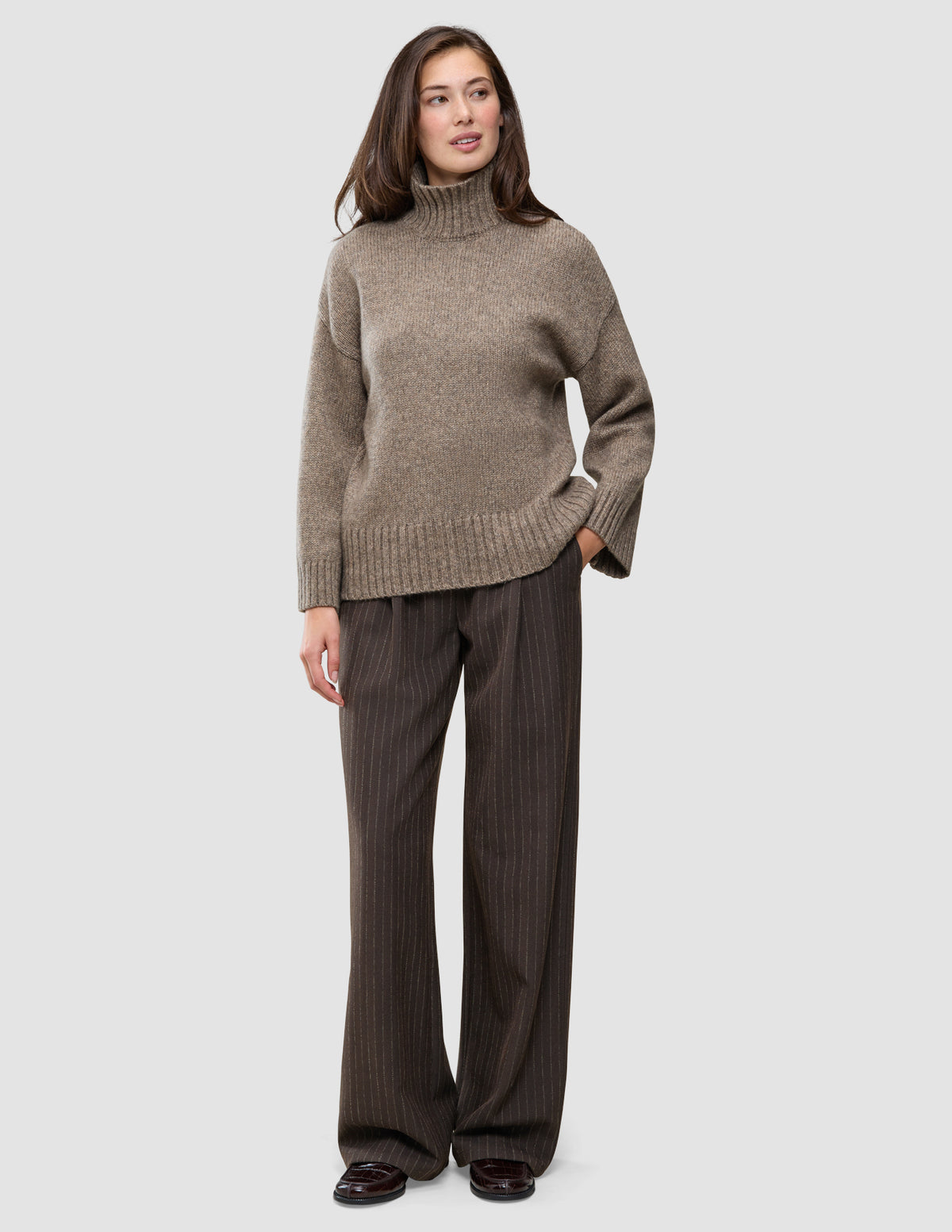 En person kledd i en tykk, myk ullstrikket turtleneck for kvinner i fargen taupe melange og brune bukser med smale striper.