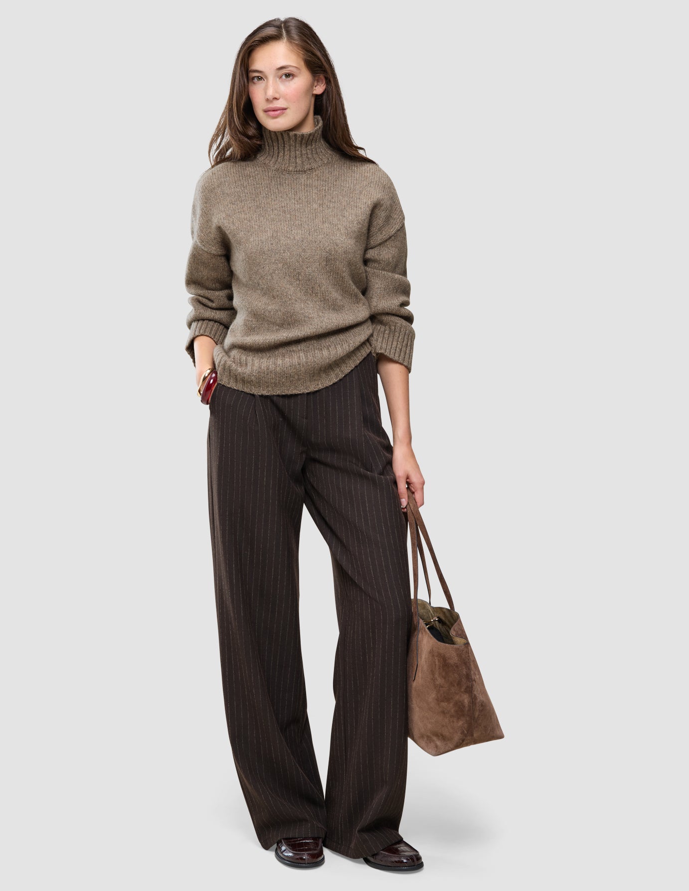 En kvinne kledd i en tykk, myk ullstrikket turtleneck for kvinner i fargen taupe melange og mørkebrune bukser med smale striper, som bærer en totebag.
