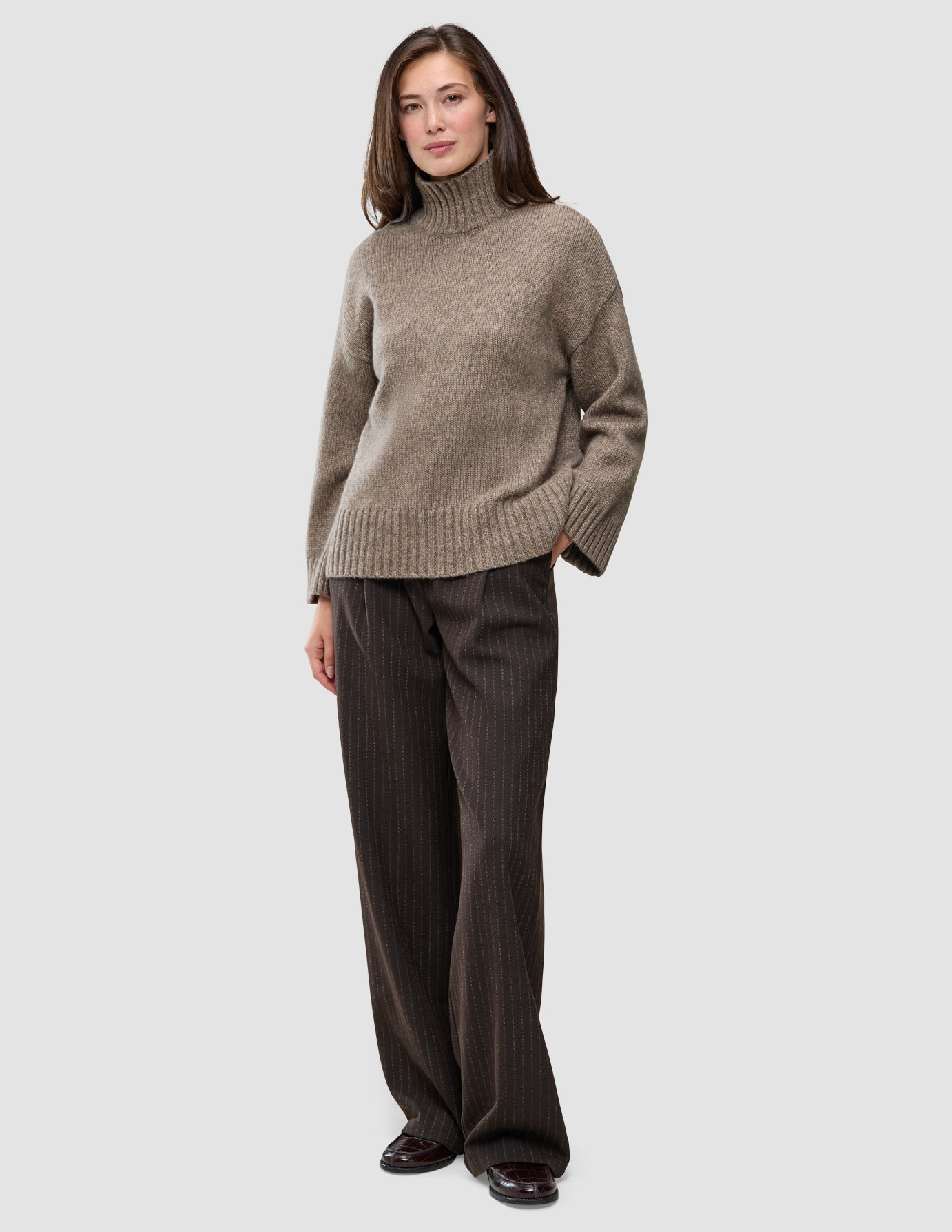 En kvinne kledd i en tykk, myk ullstrikket turtleneck for kvinner i fargen taupe melange og bukser med vid benvidde og smale striper.