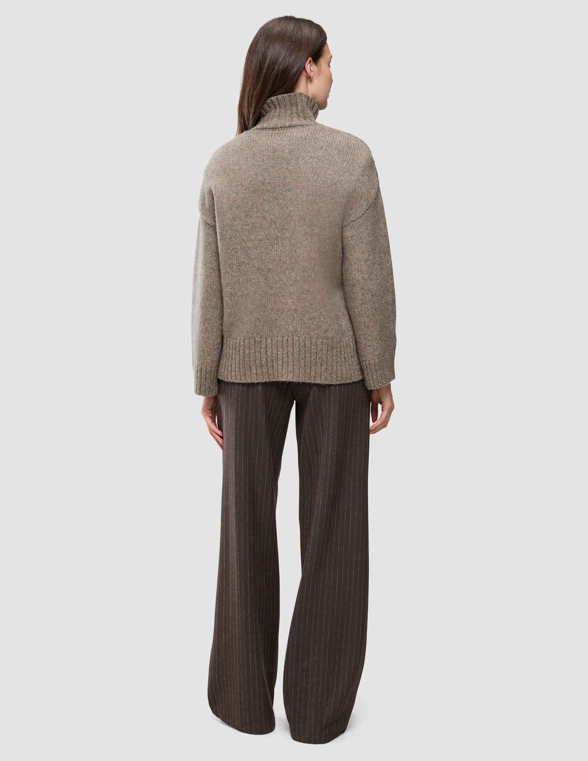 Baksiden av en tykk, myk ullstrikket turtleneck for damer i taupe-melange, kombinert med mørke bukser med smale striper.