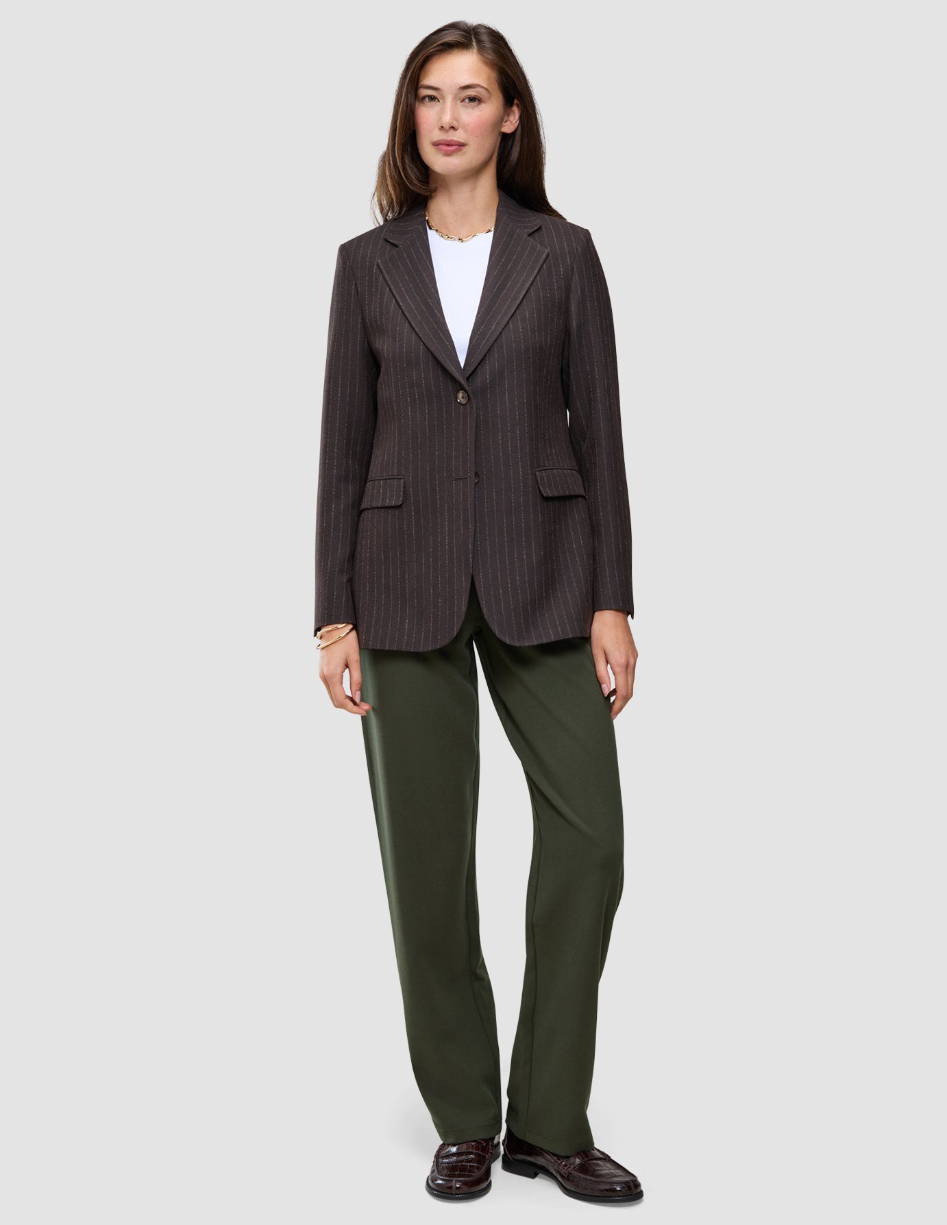 En kvinne i en brun blazer med smale striper, hvit T-skjorte, Women GO2 Pants Straight i fargen Dusty Olive og mørke loafers.