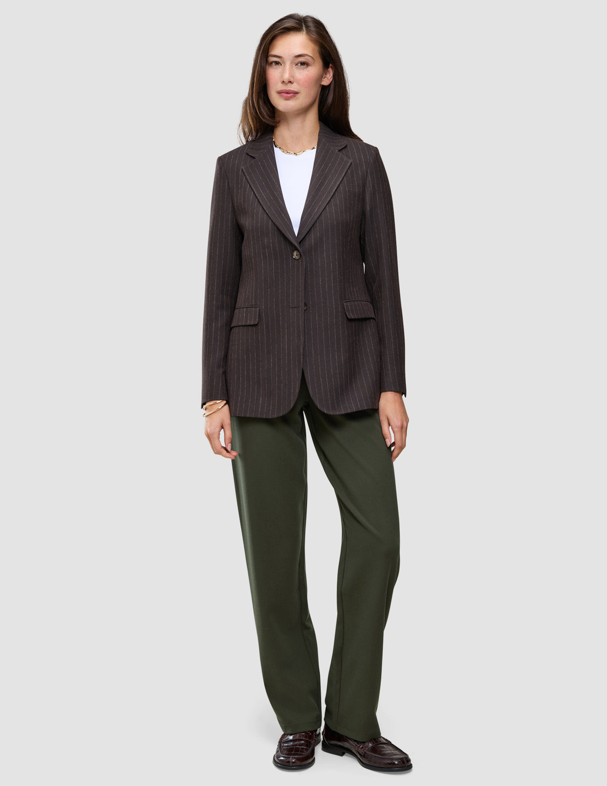 En kvinne i en brun blazer med smale striper, hvit T-skjorte, Women GO2 Pants Straight i fargen Dusty Olive og mørke loafers.
