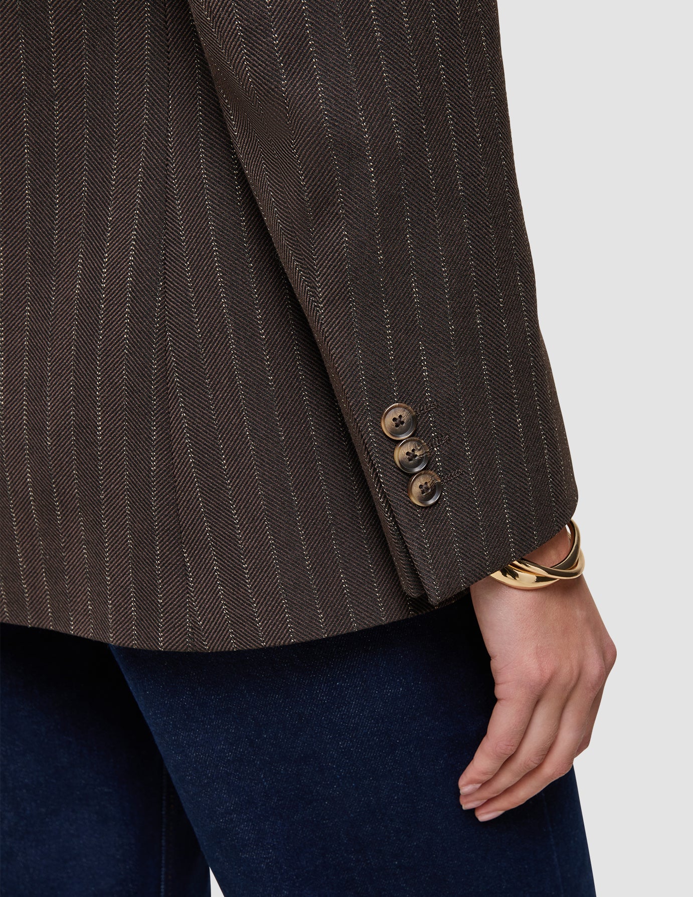 Nærbilde av en brun Women Serene Blazer Regular Walnut Herringbone-jakke med tre knapper på ermet og et gullarmbånd.