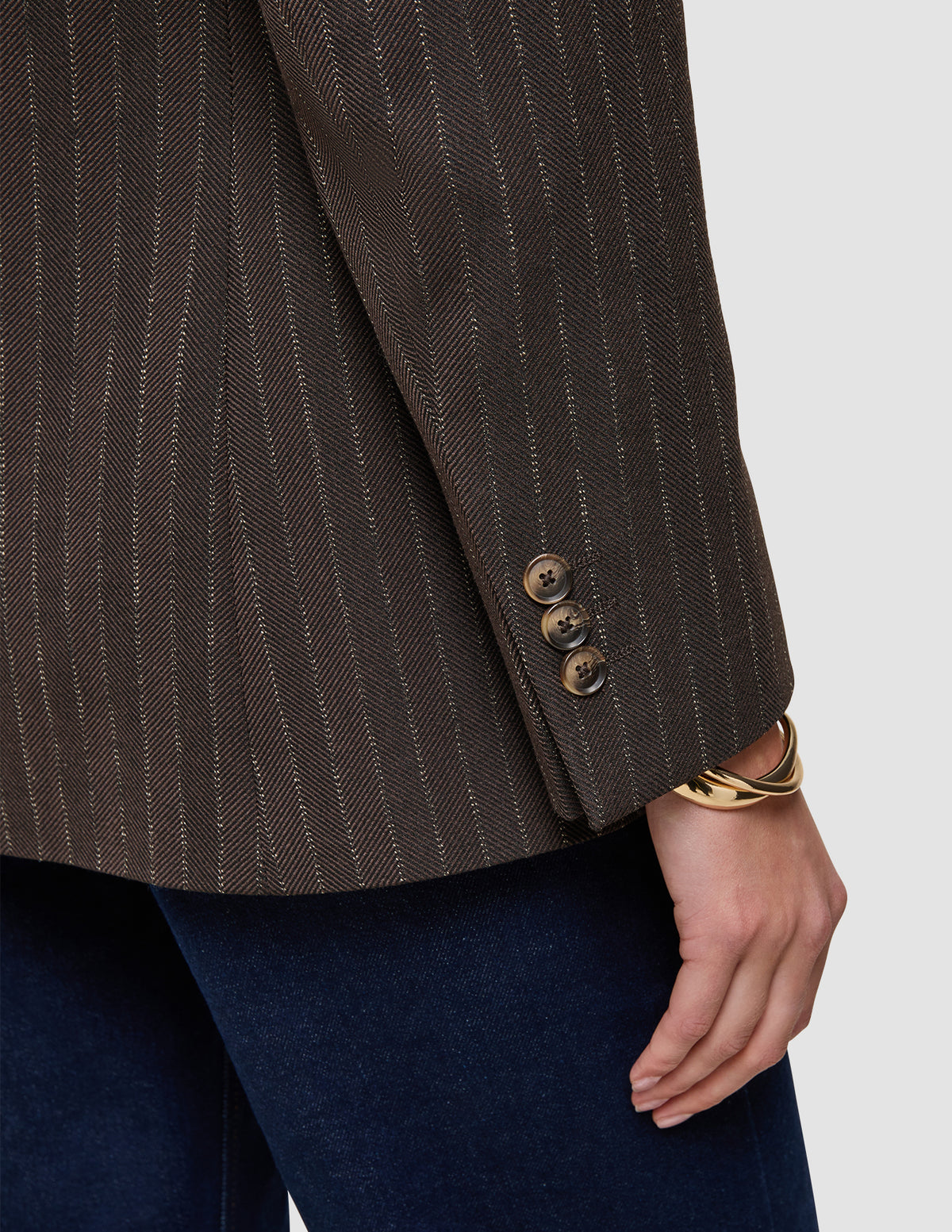 Nærbilde av en brun Women Serene Blazer Regular Walnut Herringbone-jakke med tre knapper på ermet og et gullarmbånd.